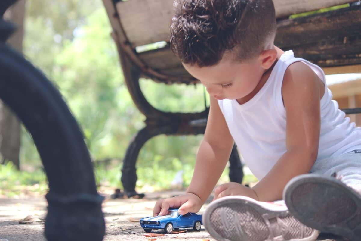 boy in white tank top playing blue coupe die cast near brown wooden bench chair during daytime