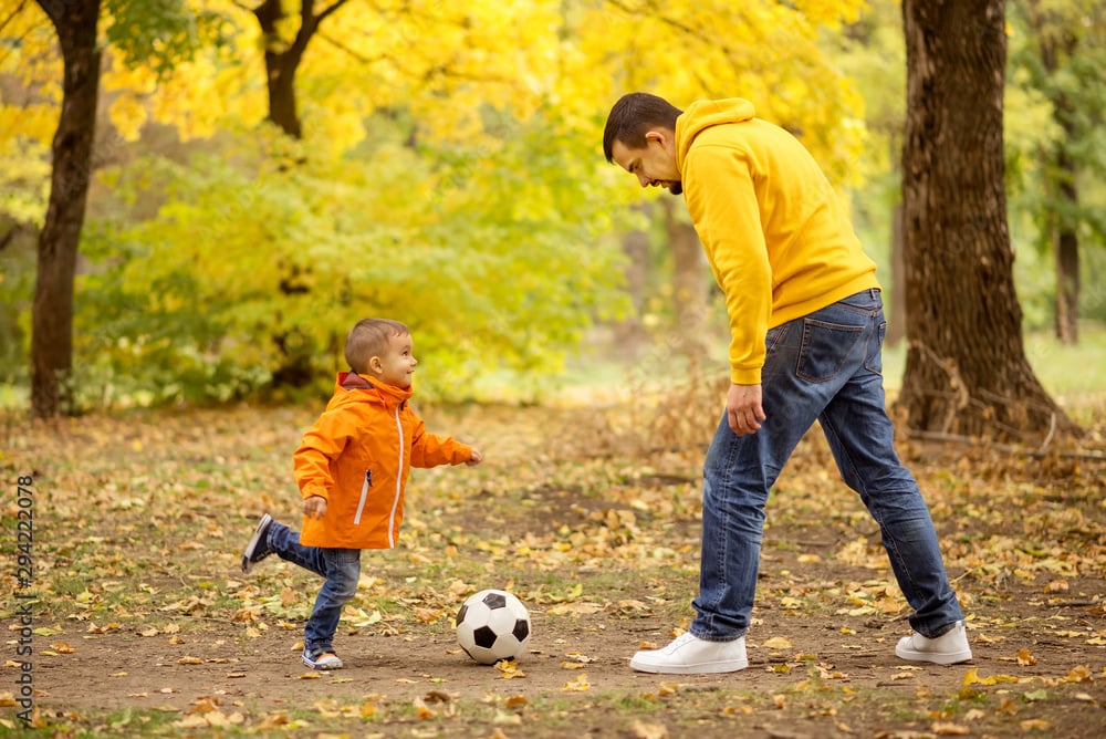 Father and little toddler son having fun in autumn park: dad and child playing football outdoors, kid raised leg ready to kick soccer ball. Active happy family and healthy parenting concept