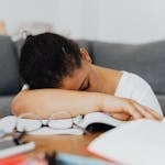 A student falls asleep while studying at home, leaning on an open book with glasses visible.