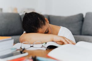 A student falls asleep while studying at home, leaning on an open book with glasses visible.